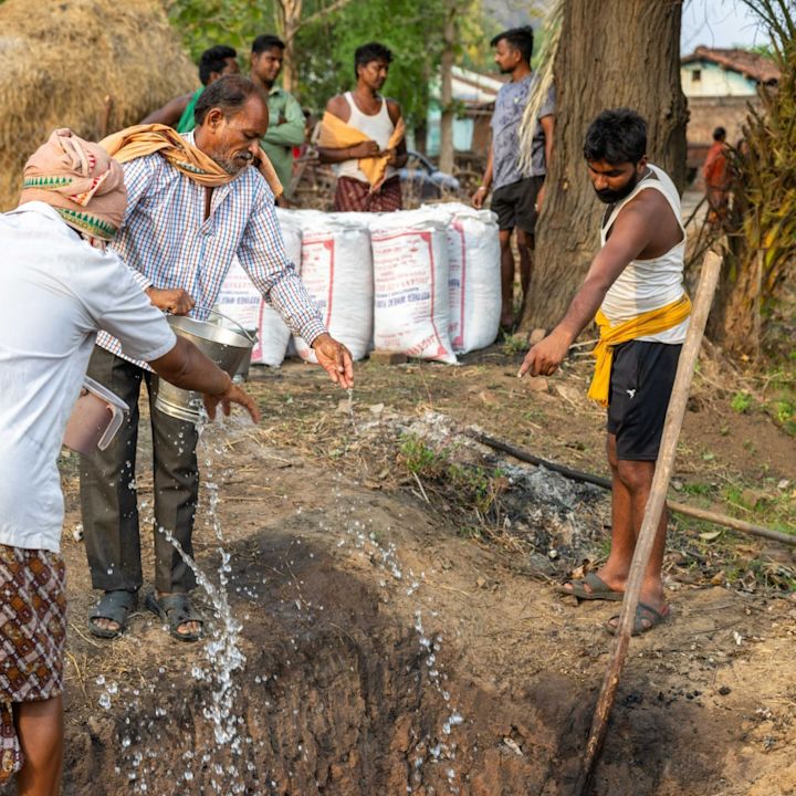 Menn som heller vann fra bøtter ned i en grøft utendørs, med flere menn som står i bakgrunnen nær store sekker og trær.